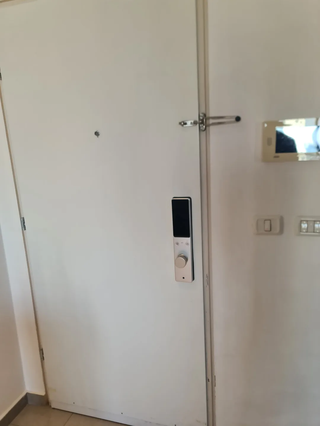 הצד הפנימי של מנעול חכם Amsterdam כסף על דלת לבנה, לצד מסך אינטרקום בכניסת הבית