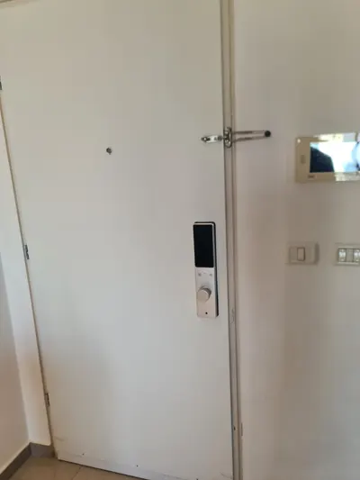 הצד הפנימי של מנעול חכם Amsterdam כסף על דלת לבנה, לצד מסך אינטרקום בכניסת הבית